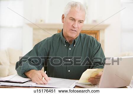 Stock Image - Man in dining room with laptop and paperwork looking unhappy. Fotosearch