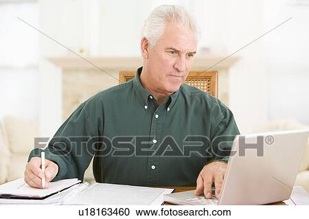 Stock Image - Man in dining room with laptop and paperwork. Fotosearch