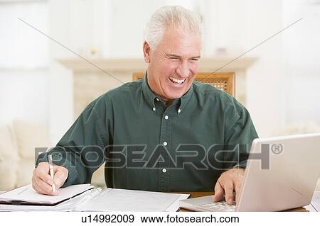 Stock Photo - Man in dining room with laptop and paperwork smiling. Fotosearch