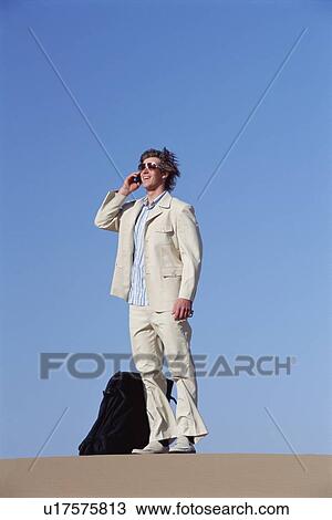Stock Image - Man outdoors with suitcase using cellular phone. Fotosearch