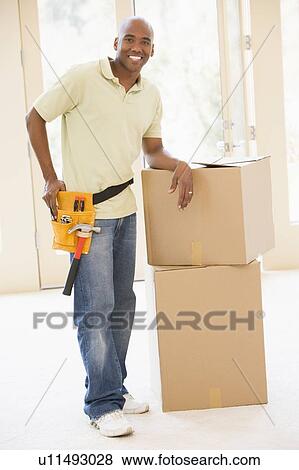 Stock Photo - Man wearing tool belt standing by boxes in new home smiling. Fotosearch
