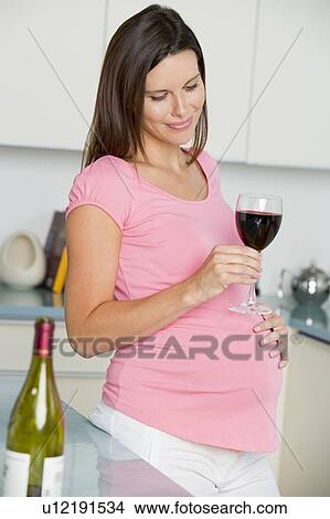 Picture - Pregnant woman in kitchen with glass of red wine smiling. Fotosearch