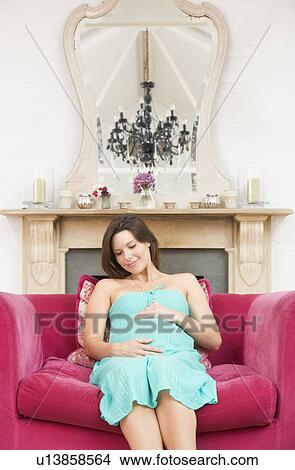 Picture - Pregnant woman sitting in living room smiling. Fotosearch