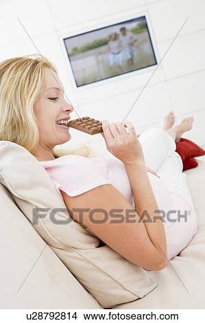 Picture - Pregnant woman watching television and eating chocolate smiling. Fotosearch