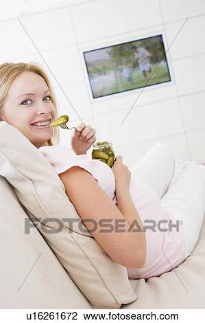 Stock Image - Pregnant woman watching television and eating pickles smiling. Fotosearch