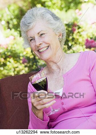 Picture - Senior woman sitting outdoors on a chair with a glass of red wine. Fotosearch