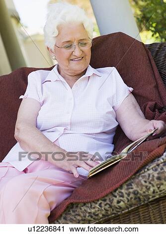 Stock Image - Senior woman sitting outdoors on a chair reading a book. Fotosearch