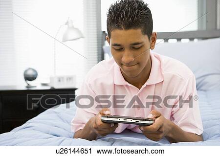 Stock Image - Teenage Boy Lying On Bed Playing Video Game. Fotosearch