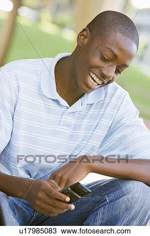 Stock Image - Teenage Boy Sitting Outdoors Using Mobile Phone. Fotosearch