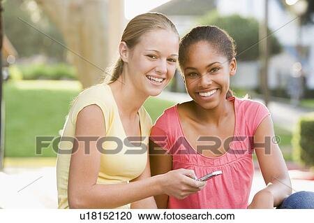 Stock Image - Teenage Girls Sitting Outside Playing With Mobile Phone. Fotosearch