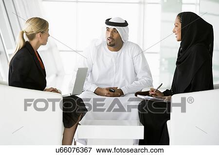 Stock Photo - Three businesspeople indoors with laptop talking and smiling (high key/selective focus). Fotosearch