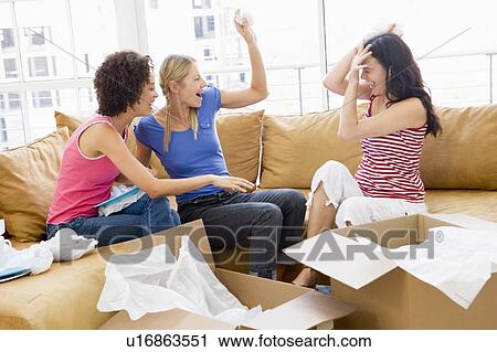 Stock Image - Three girl friends playfully unpacking boxes in new home smiling. Fotosearch