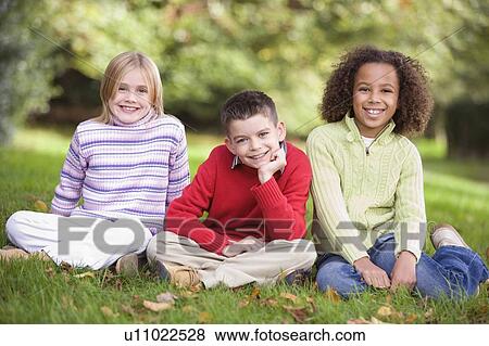 Three young children sitting outdoors in park smiling (selective focus) View Large Photo Image Stock Photo - Three young children sitting outdoors in park smiling (selective focus). Fotosearch