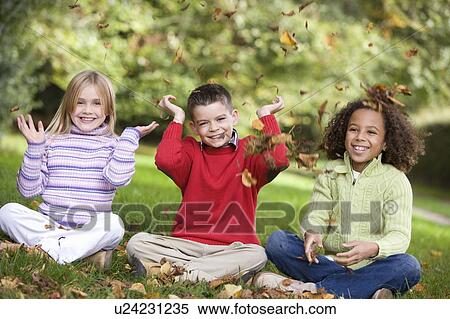 Stock Photography - Three young children sitting outdoors in park throwing leaves in air and smiling (selective focus). Fotosearch