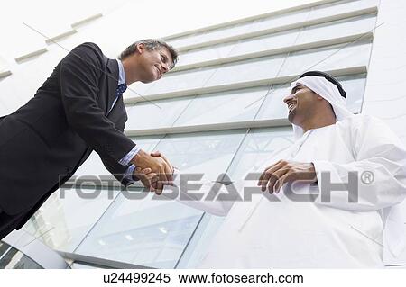 Stock Photography - Two businessman outdoors by building shaking hands and smiling (high key/selective focus). Fotosearch