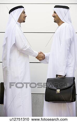 Stock Image - Two businessmen outdoors shaking hands and smiling. Fotosearch