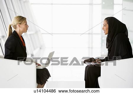 Stock Photograph - Two businesswomen indoors with laptop talking and smiling (high key/selective focus). Fotosearch