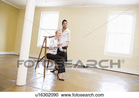 Stock Image - Two women in empty space with ladder holding paper and smiling. Fotosearch