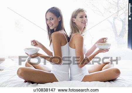Stock Photo - Two women sitting on bed eating cereal smiling. Fotosearch