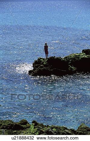 Stock Image - Woman outdoors standing on large rocks Oceanside (far away). Fotosearch