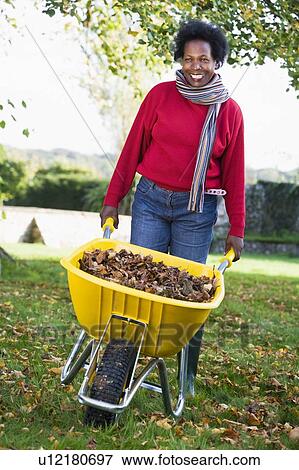 Stock Photo - Woman outdoors with leaves in wheelbarrow smiling (selective focus). Fotosearch