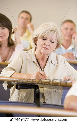 Stock Photo - Woman sitting in adult classroom taking notes with students in background (selective focus). Fotosearch