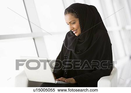 Stock Photography - Woman sitting indoors with laptop smiling (high key/selective focus). Fotosearch