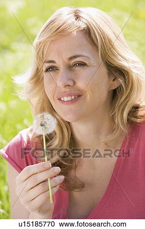 Stock Image - Woman sitting outdoors holding dandelion head smiling. Fotosearch