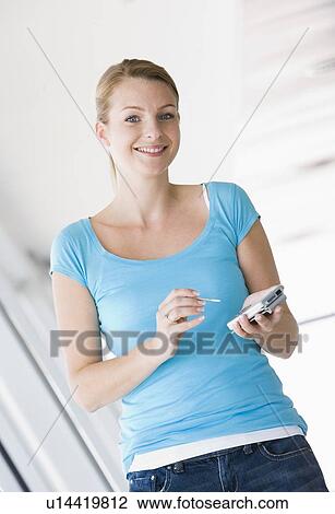 Stock Image - Woman standing in corridor using personal digital assistant smiling. Fotosearch