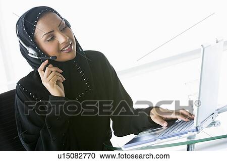 Stock Image - Woman wearing headset with laptop smiling (high key/selective focus). Fotosearch