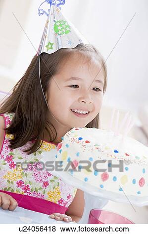 Stock Photo - Young girl wearing party hat looking at birthday cake smiling. Fotosearch