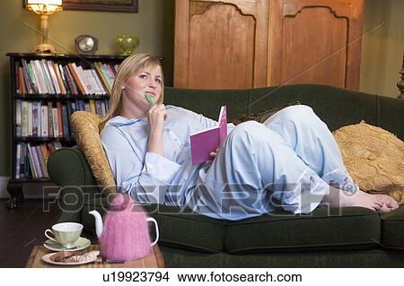 Picture - Young woman lying on sofa at home writing in her diary. Fotosearch