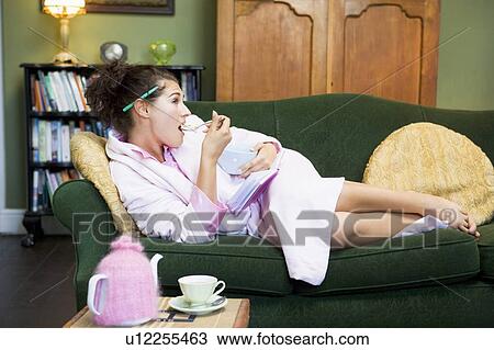 Stock Image - Young woman lying on sofa at home eating a sweet treat. Fotosearch