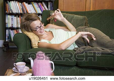 Stock Image - Young woman lying on sofa at home eating chocolate. Fotosearch
