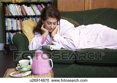 Stock Image - Young woman lying on sofa at home watching telephone. Fotosearch