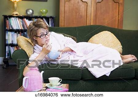 Stock Photograph - Young woman lying on sofa at home eating a sweet treat. Fotosearch
