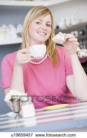 Stock Photo - Young woman sitting at a table drinking tea and eating a sweet treat. Fotosearch
