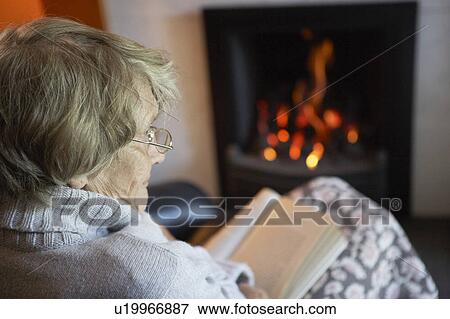 Stock Photo - Senior Woman Reading Book By Fire At Home. Fotosearch