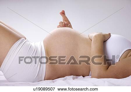 Stock Photo - Midsection of a pregnant young woman lying on her back, fingers crossed. Fotosearch