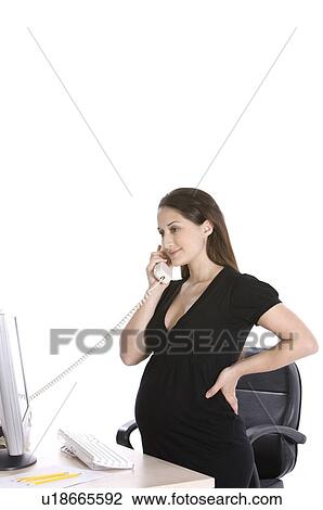 Stock Image - Pregnant woman on the phone, standing in front of a desktop PC. Fotosearch