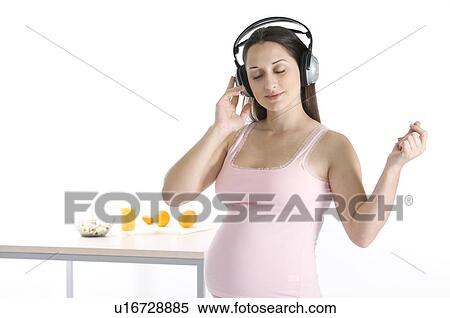 Stock Photography - Pregnant woman with headphones on, standing next to a table with food on it. Fotosearch
