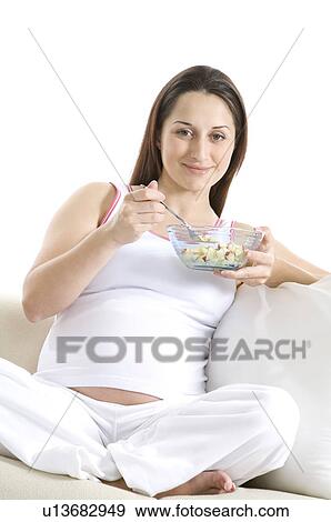 Stock Photo - Pregnant young woman eating from a bowl, sitting on sofa. Fotosearch