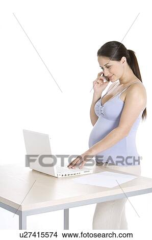 Picture - Pregnant young woman on the phone, looking at a laptop, standing. Fotosearch