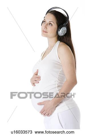 Stock Image - Pregnant young woman wearing headphones, hands around the abdomen, looking up. Fotosearch