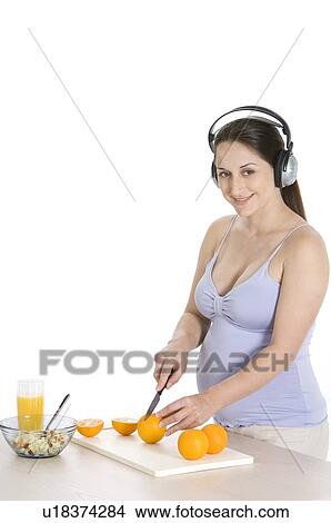 Picture - Pregnant young woman with headphones on, cutting an orange. Fotosearch