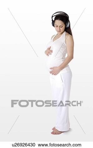 Stock Image - Pregnant young woman with headphones on, touching abdomen lightly, standing. Fotosearch