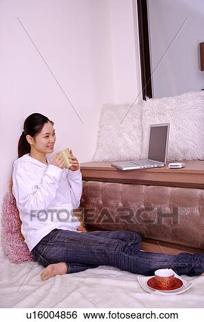 Stock Photograph - Young woman holding tea cup watching her laptop. Fotosearch