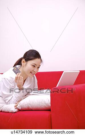 Young woman lying on a sofa, holding a credit card in her hand and using a laptop View Large Photo Image Stock Photo - Young woman lying on a sofa, holding a credit card in her hand and using a laptop. Fotosearch
