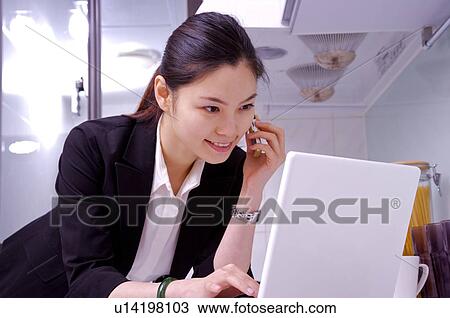 Stock Image - Young woman on the phone at kitchen counter using laptop. Fotosearch