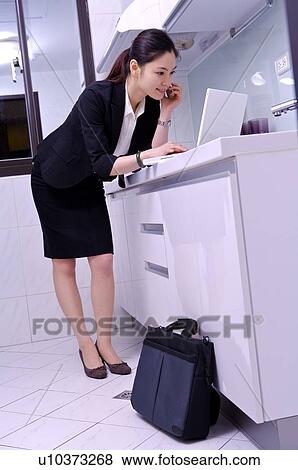Stock Photo - Young woman on the phone at kitchen table using laptop computer. Fotosearch